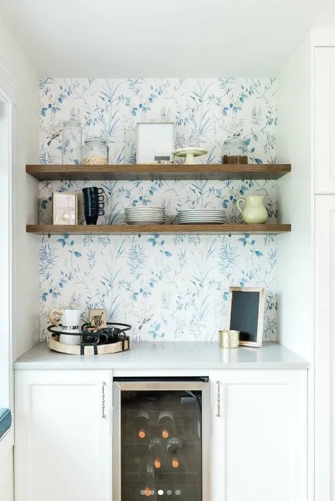 Compact coffee bar with blue floral wallpaper, wooden floating shelves, and white lower cabinets